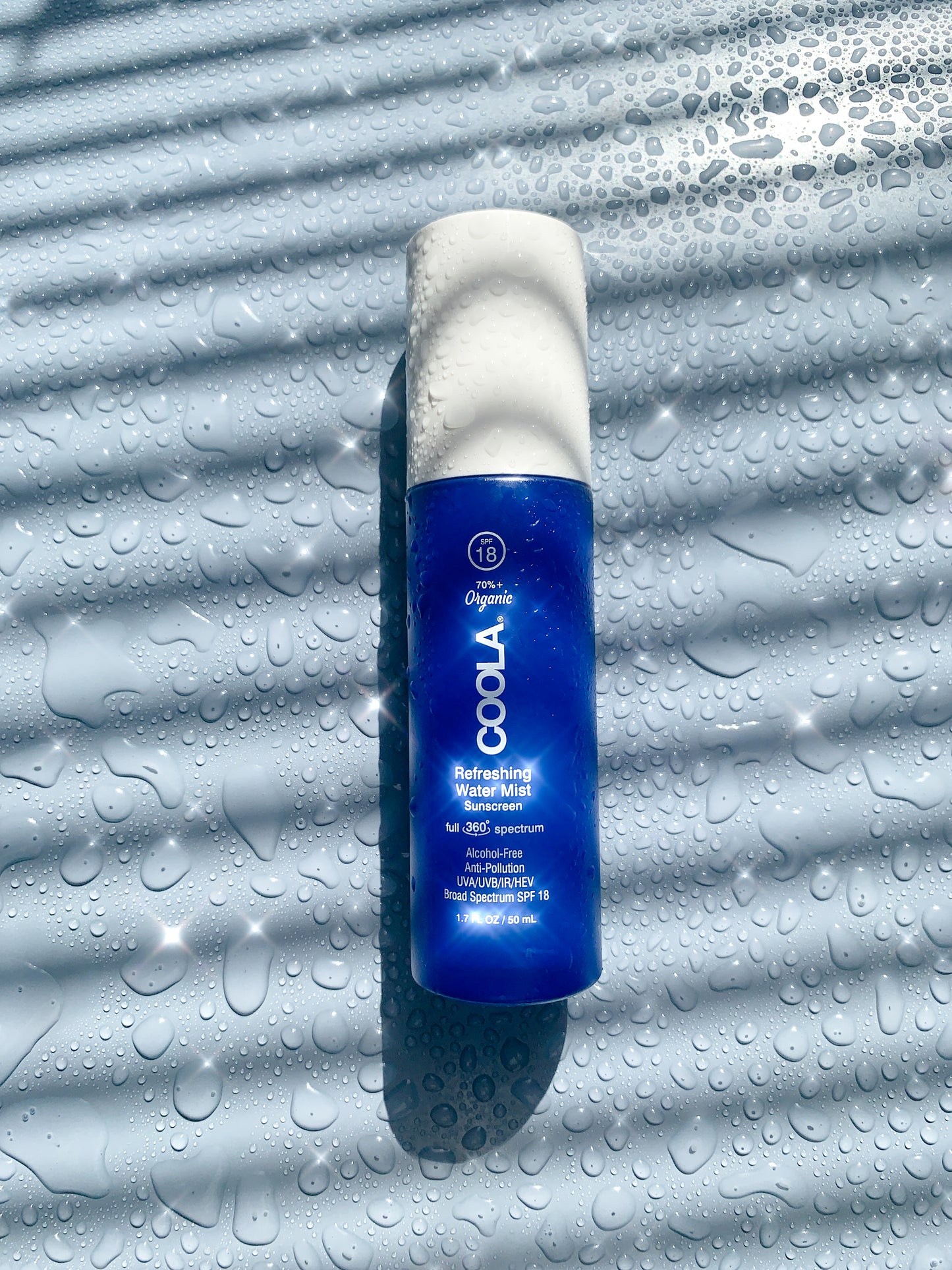 Brume d’eau rafraîchissante Coola SPF18 posée sur une surface texturée avec gouttelettes, idéale pour une hydratation rapide et une protection solaire légère.