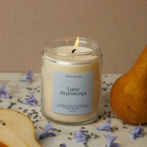 Chandelle Lueur du Printemps de Selv Rituel, allumée et entourée de poires, lavande et fleurs violettes, pour une atmosphère fraîche et florale.