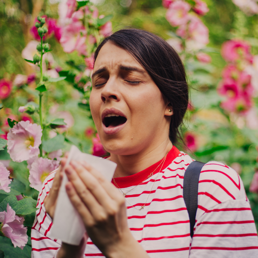 Une femme éternuant dans un jardin fleuri, symbole des allergies saisonnières souvent déclenchées par le pollen, apaisées par des solutions naturelles.
