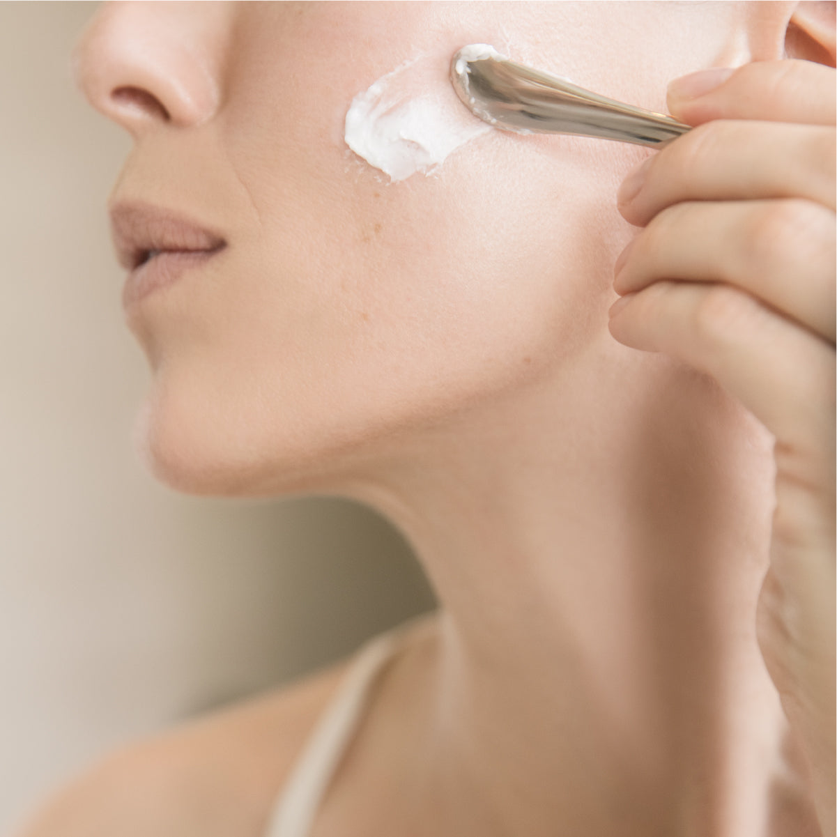 Application d’une crème anti-âge sur la joue avec une spatule métallique, mettant en valeur la texture riche de la EGF Power Cream de Bioeffect.