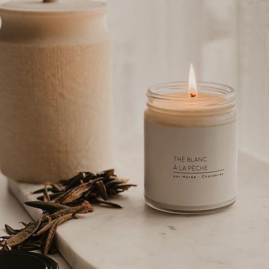 Chandelle The Blanc a la Peche allumee, accompagnee de feuilles de the sechees sur une table en marbre, parfaite pour une atmosphère relaxante.