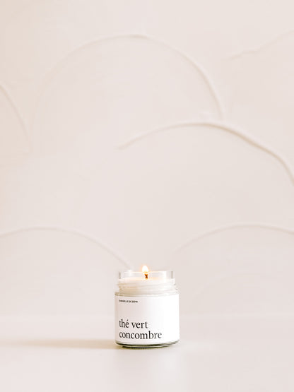 Une chandelle de soya the vert concombre illuminee sur une surface neutre avec un mur clair en arriere-plan.