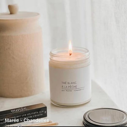 Chandelle The Blanc a la Peche allumee, posée sur une table en marbre avec un couvercle noir et des accessoires en bois, dégageant une ambiance apaisante.