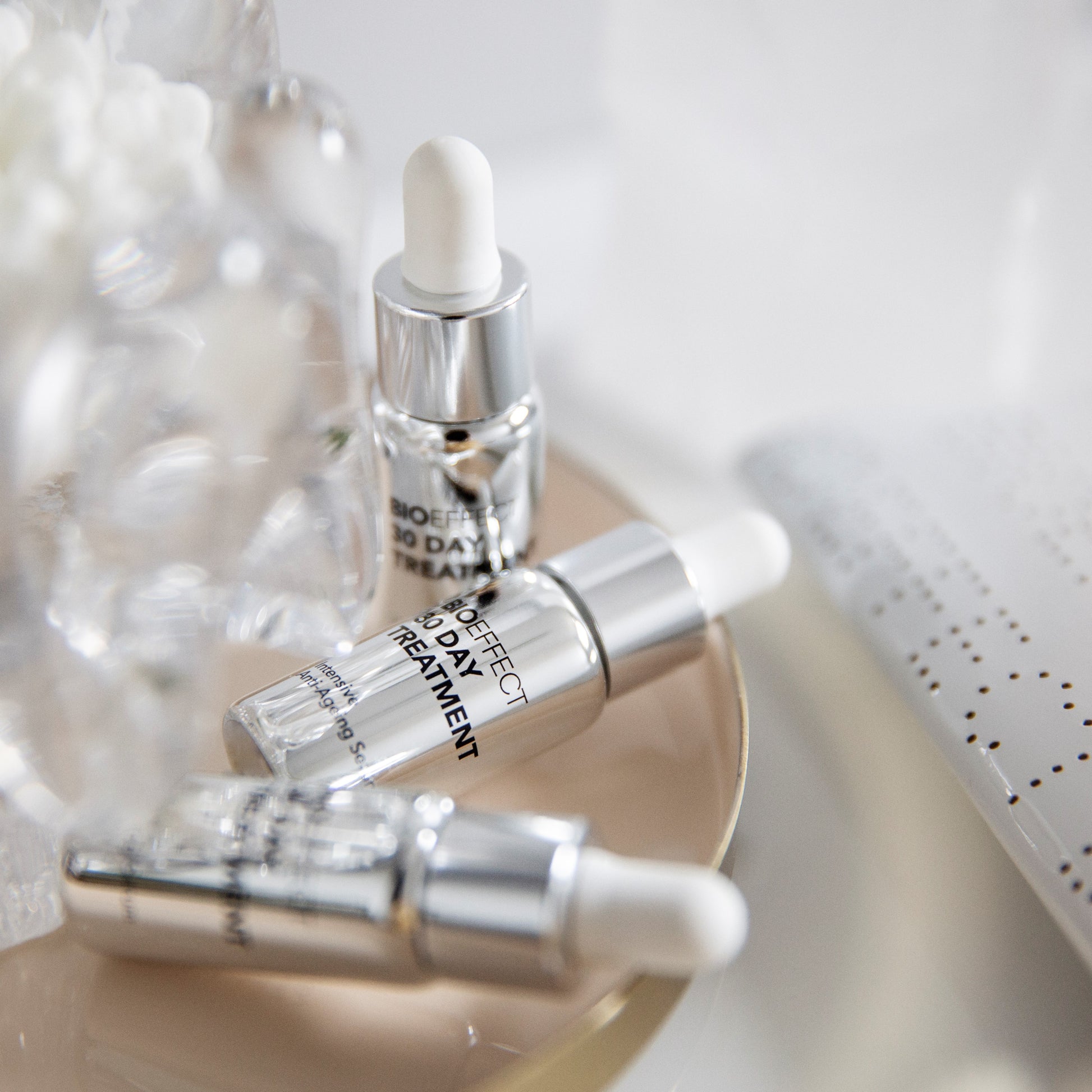 Trois flacons du traitement Bioeffect 30 Day posés sur une assiette beige, entourés d'éléments décoratifs flous, ambiance douce et raffinée.