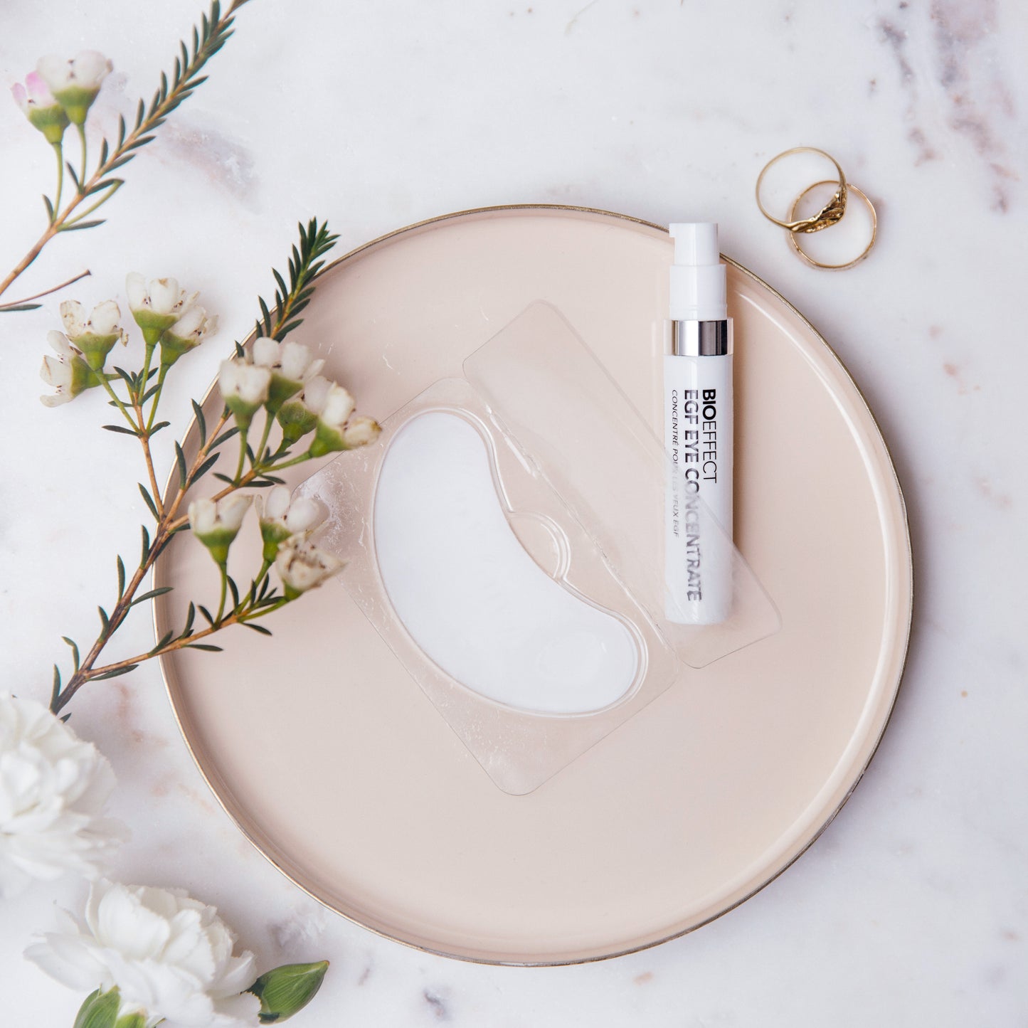 Sérum contour des yeux Bioeffect et patch hydrogel disposés sur une assiette rose, entourés de fleurs naturelles et d'anneaux dorés, ambiance douce et raffinée.