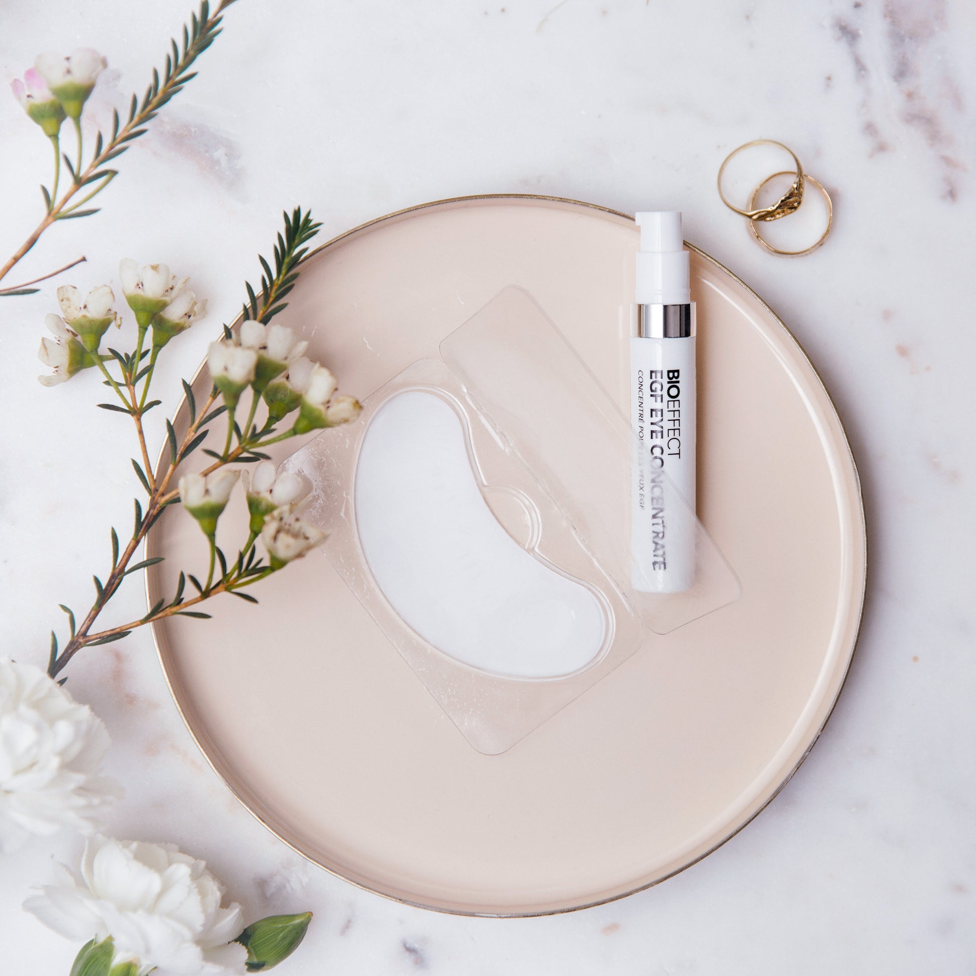 Sérum contour des yeux Bioeffect et patch hydrogel disposés sur une assiette rose, entourés de fleurs naturelles et d'anneaux dorés, ambiance douce et raffinée.