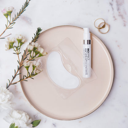Sérum contour des yeux Bioeffect et patch hydrogel disposés sur une assiette rose, entourés de fleurs naturelles et d'anneaux dorés, ambiance douce et raffinée.